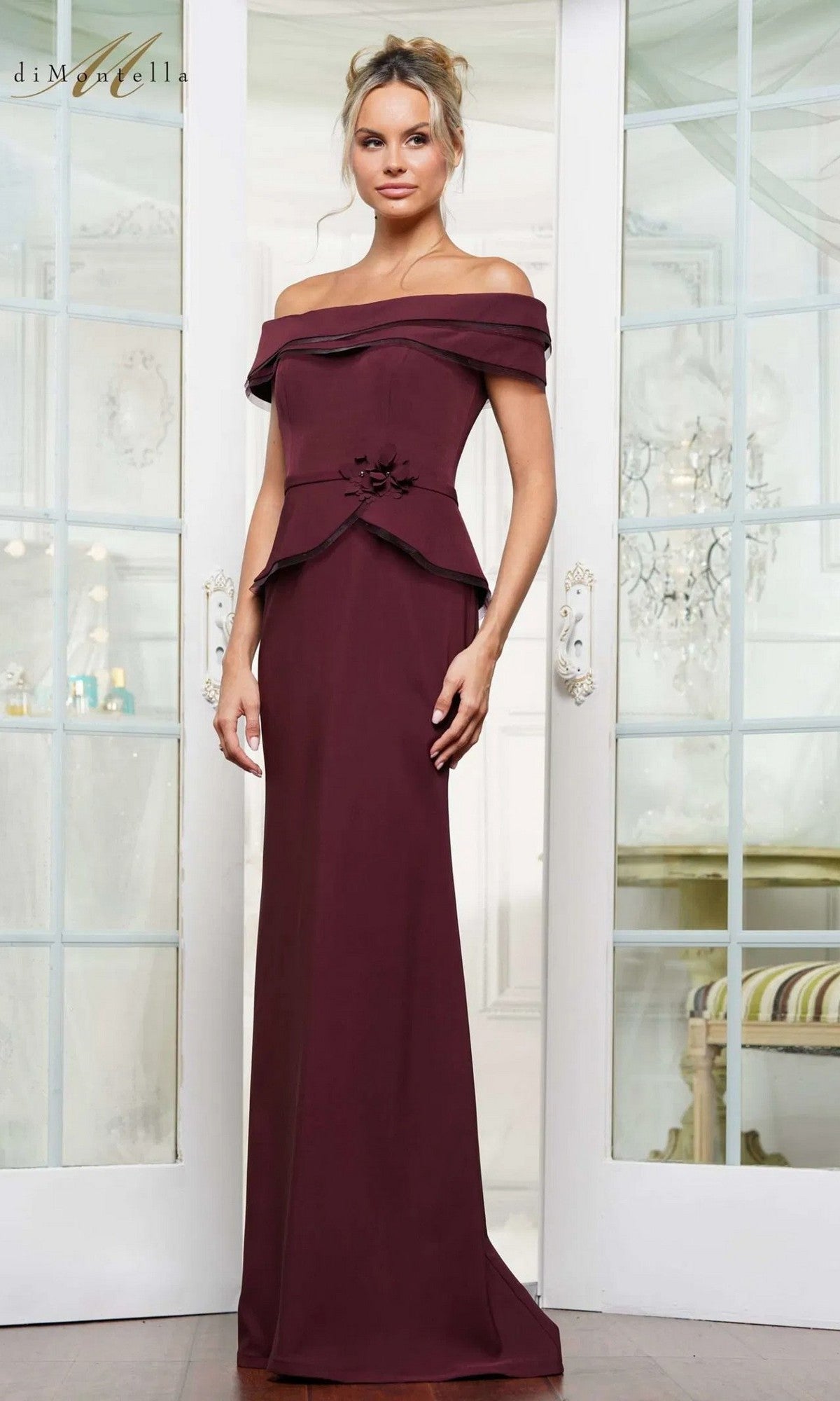 A woman in a wine-colored, floor-length Rina Di Montella RD3256 formal dress stands indoors before glass doors.
